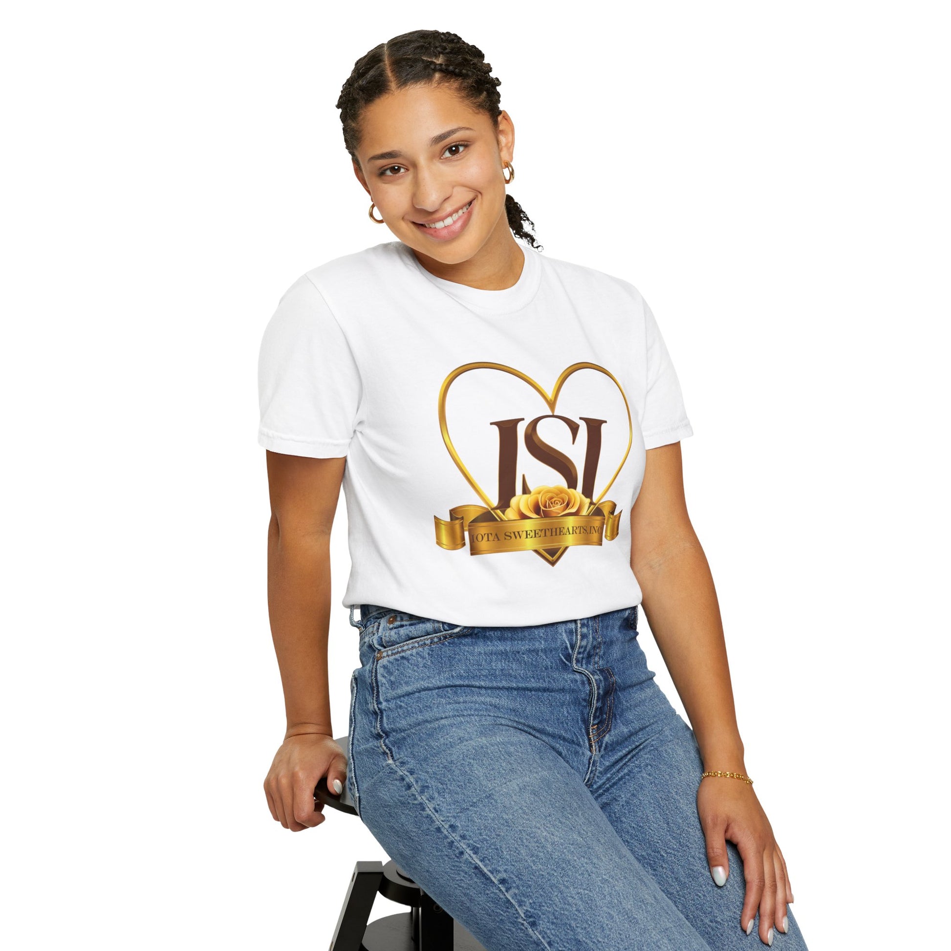 A cheerful woman with braided hair is sitting on a stool, smiling warmly while wearing a white t-shirt featuring the Iota Sweethearts, Inc. logo. The shirt design includes bold brown "ISI" letters inside a gold heart outline, with a golden ribbon and yellow rose beneath the text reading "Iota Sweethearts, Inc." She pairs the shirt with high-waisted blue jeans and accessorizes with hoop earrings and a bracelet. The background is plain white, putting full focus on the subject and t-shirt design.