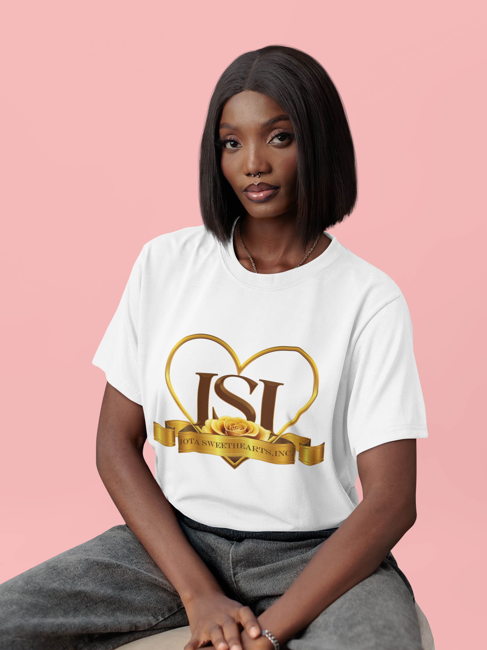 A stylish Black woman with straight shoulder-length hair is seated against a pink background, wearing a white t-shirt featuring the Iota Sweethearts, Inc. logo. The design includes a gold heart outline, bold "ISI" letters, a yellow rose, and a gold ribbon with the text "Iota Sweethearts, Inc." printed on it. She has a confident expression and is casually dressed in gray pants.