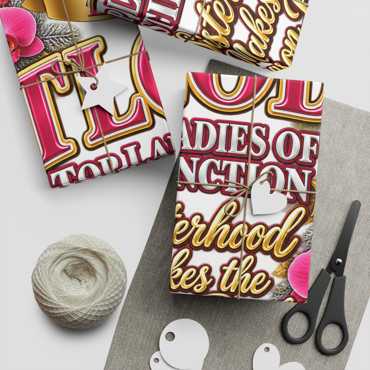 Three wrapped gift boxes with wrapped paper featuring a bold pink, white, and gold typographic design, styled on a light gray surface with twine, paper tags, a ball of thread, and black-handled scissors.