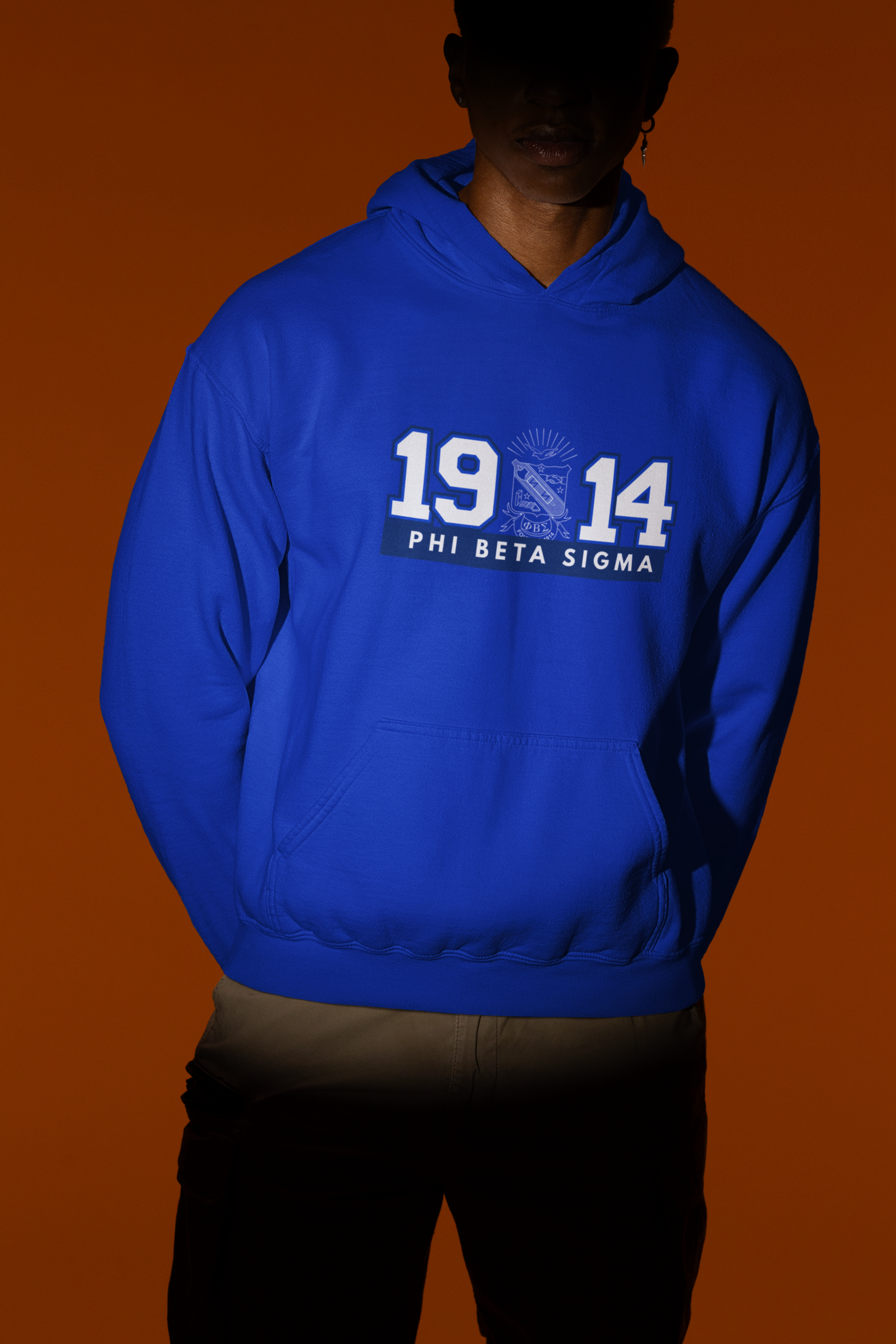 Model wearing a royal blue Phi Beta Sigma 1914 hoodie with fraternity shield crest and kangaroo pocket against a dramatic spotlight background.