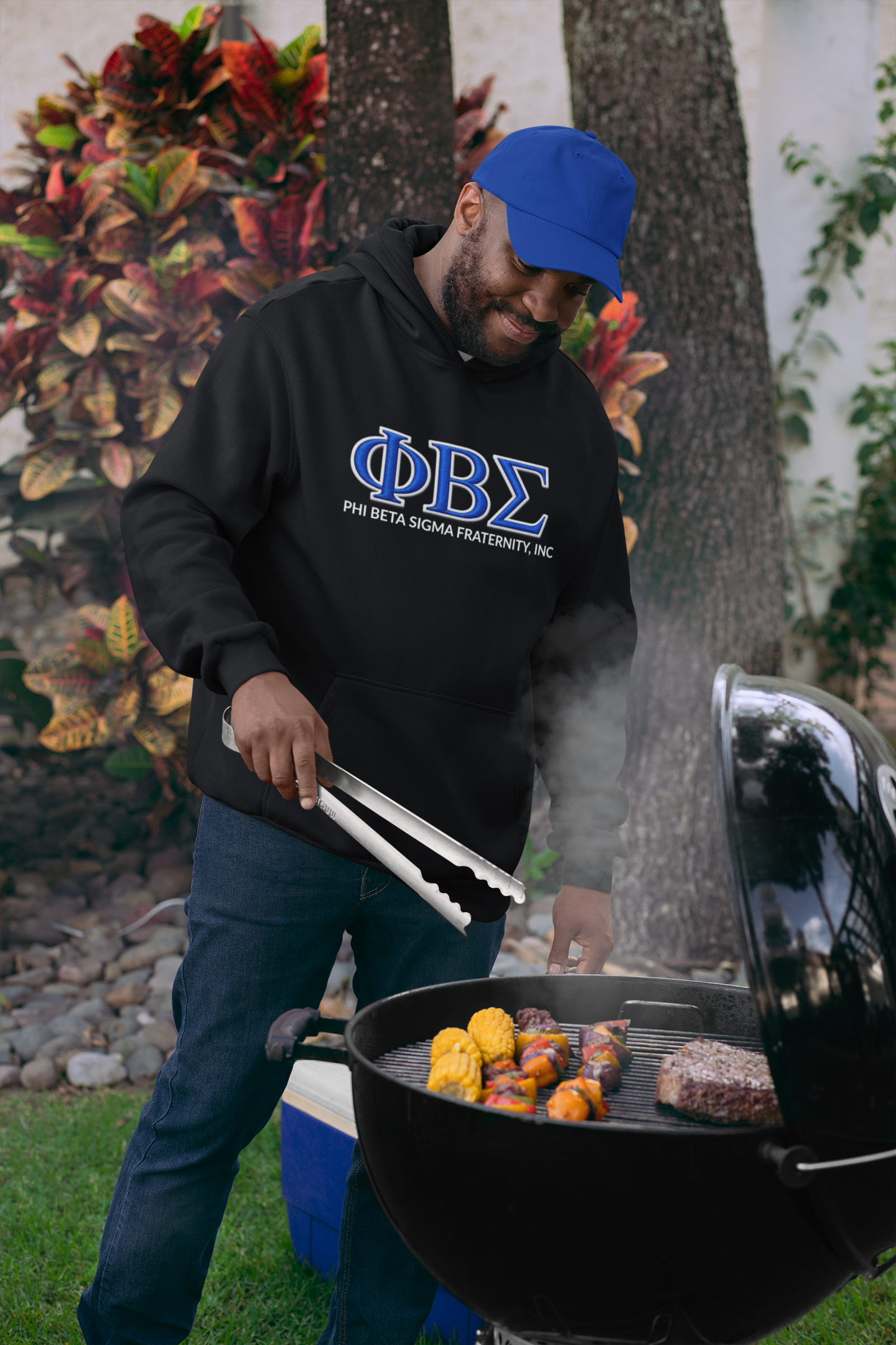 Man grilling outdoors wearing a black Pi Beta Sigma hoodie with blue text and a blue cap.