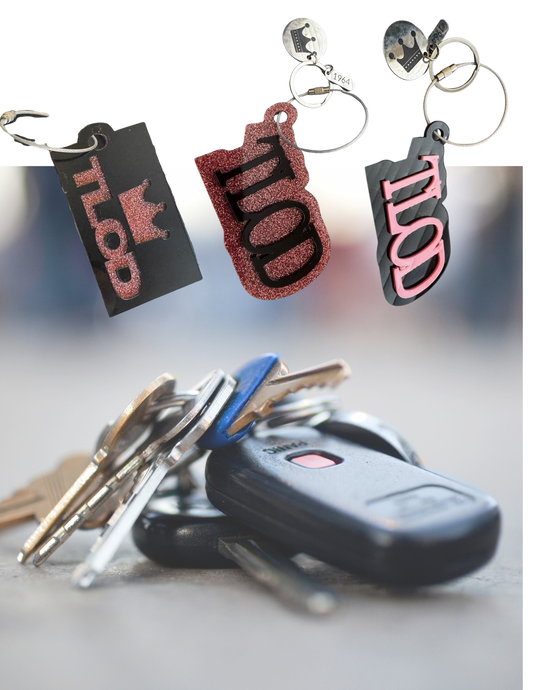 A collection of TLOD keychains in various styles, including black and pink acrylic designs with a crown charm and "1964" tag, floating above a blurred background of a set of keys with a car key fob. The keychains are positioned to showcase their unique glitter and engraved details.